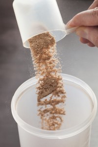 Scoop of chocolate whey isolate protein tossed into plastic white shaker, with focus on the protein in the scoop and falling protein blurred